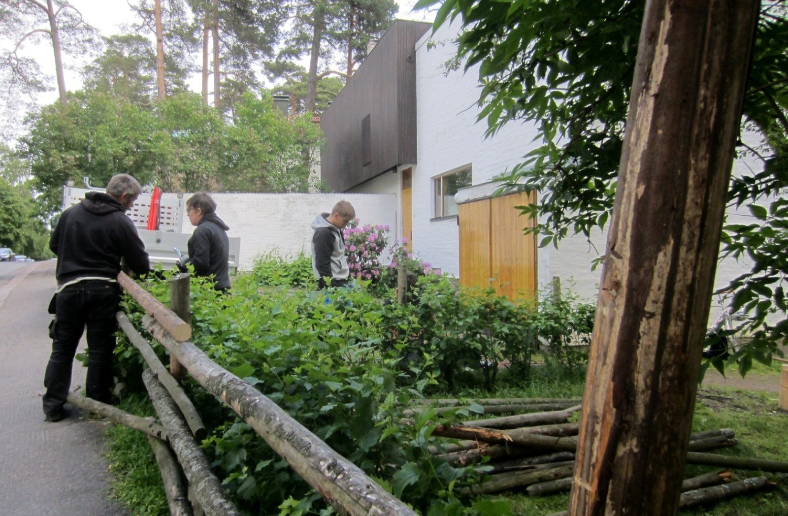 Kolme henkilöä asentaa kuorituista puukappaleista koostuvaan aitaan uusia osia. Etualalla puuosia ja taustalla vaalea rakennus.. Kuvaaja: Jonas Malmberg. Kolme henkilöä asentaa kuorituista puukappaleista koostuvaan aitaan uusia osia. Etualalla puuosia ja taustalla vaalea rakennus.. Kuvaaja: Jonas Malmberg.