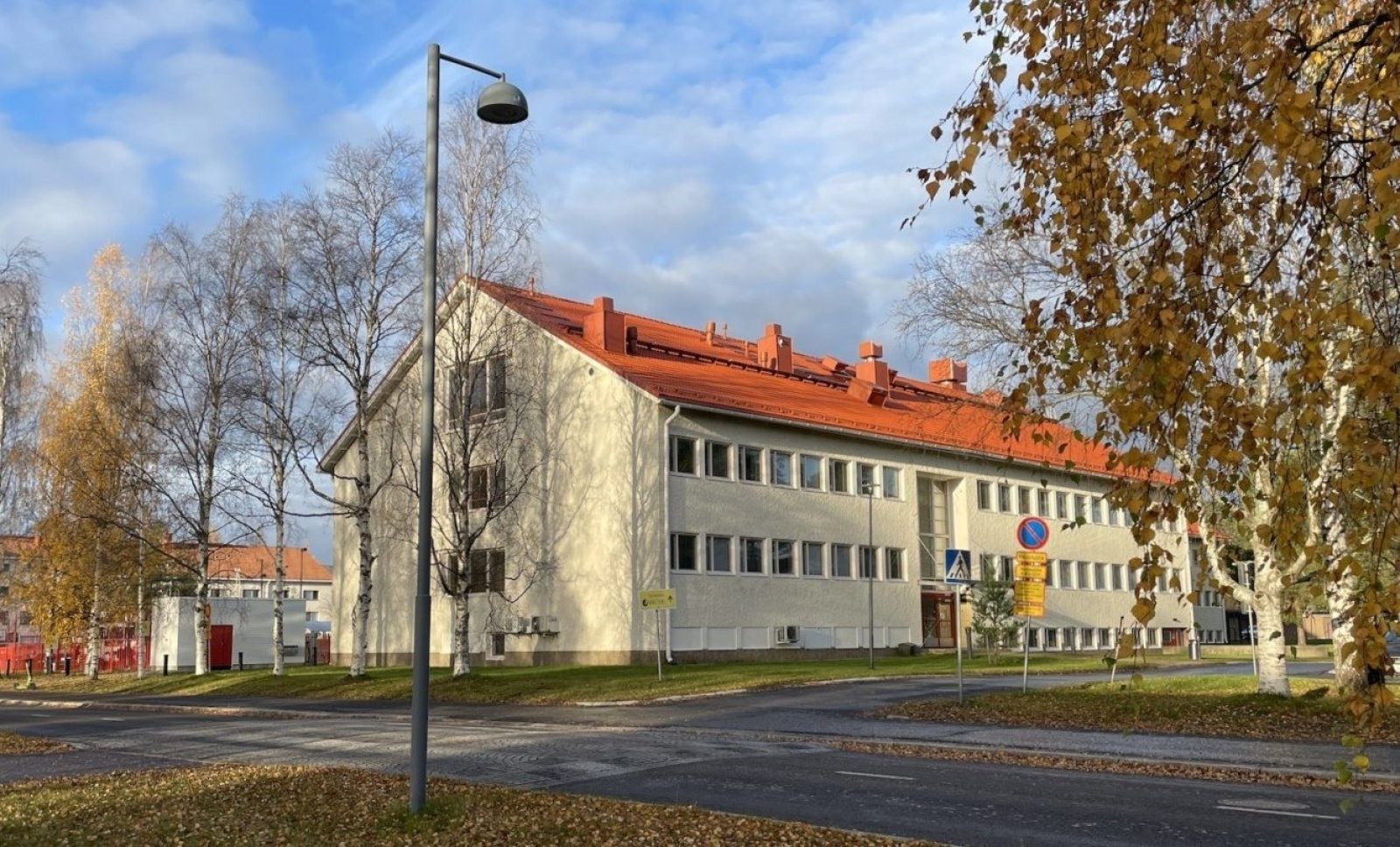 Vaaleaksi rapattu harjakattoinen rakennus, jonka edustalla on koivurivistö ja tie. Taustalla näkyy toinen samanlainen rakennus.