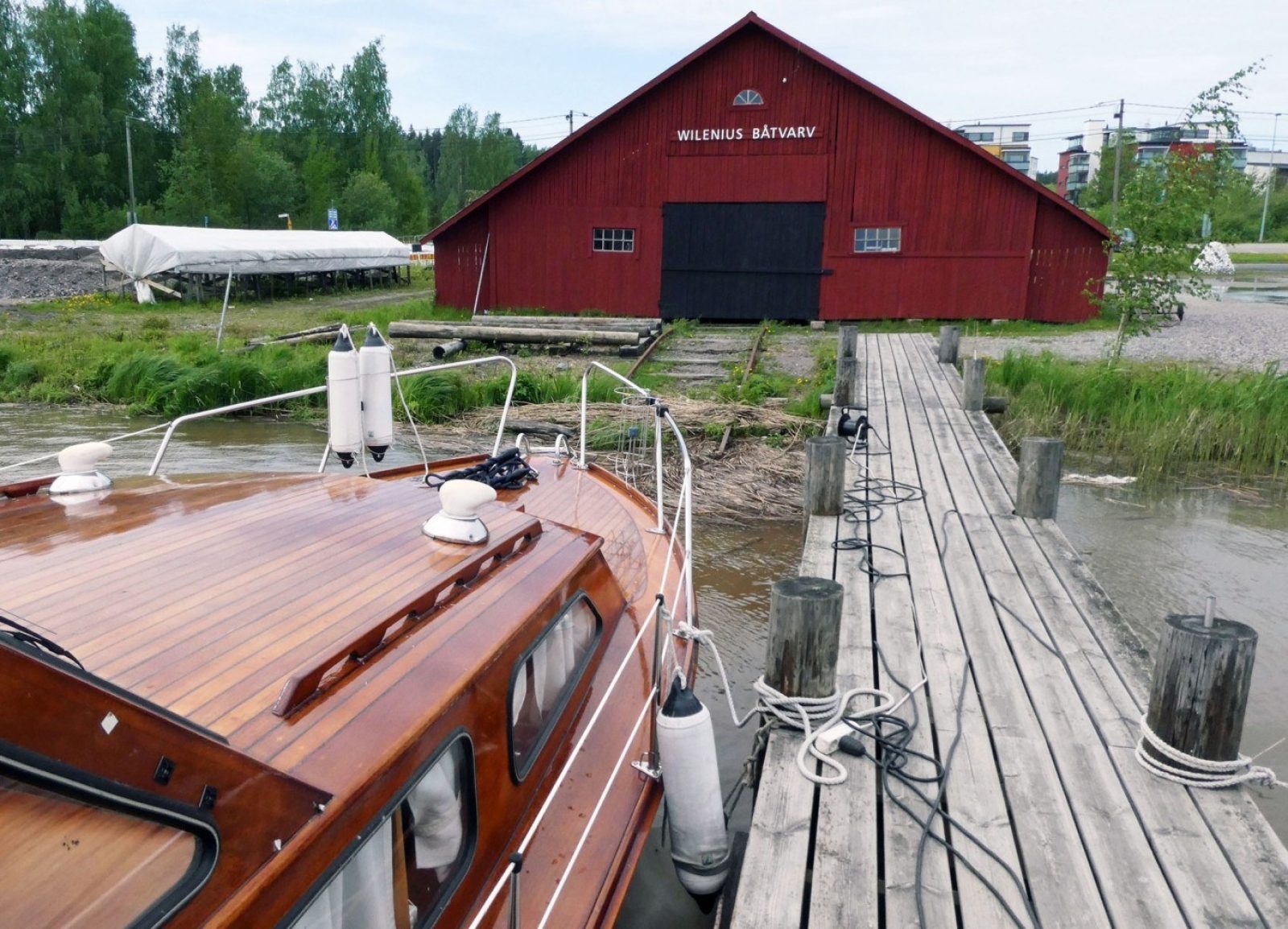 Puinen viistokattoinen veistämörakennus rannassa. Etualalla puuvene laiturissa.