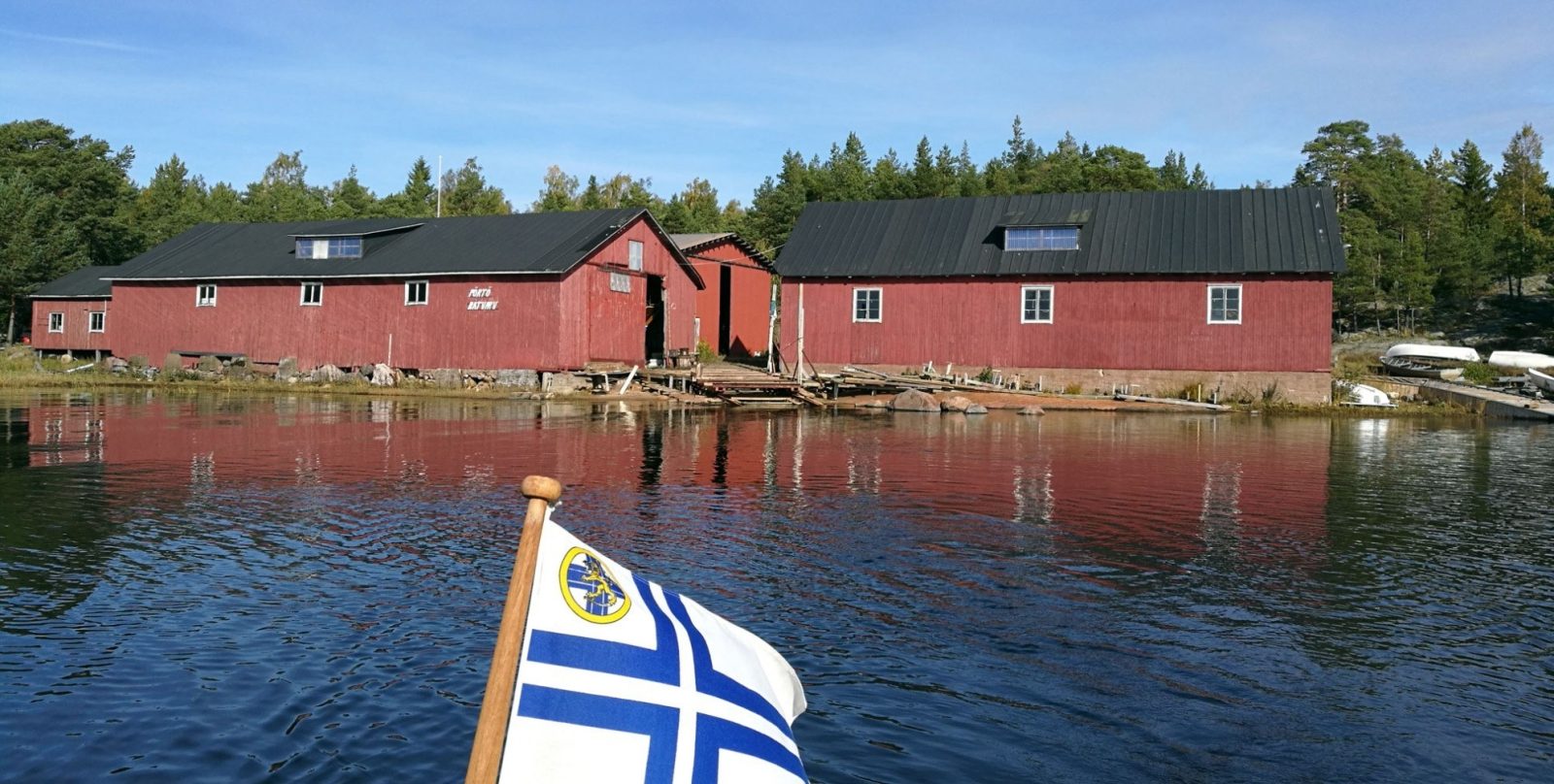 Punaisia mustakattoisia rakennuksia veden äärellä. Etualalla valkoinen riniristinen purjehdusseuran lippu.
