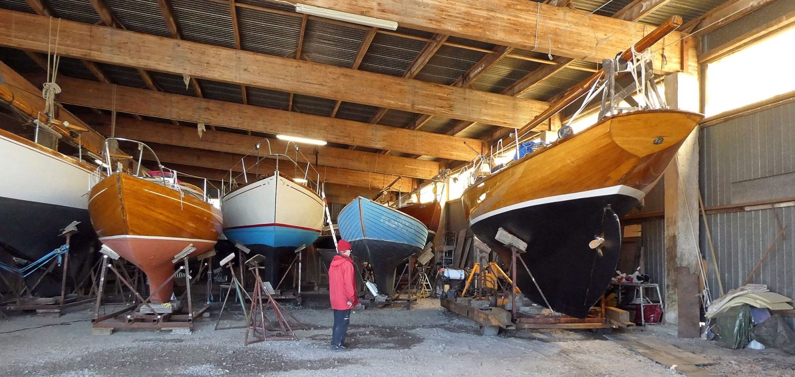 Neljä puuvenettä telineillä telakan hallissa. Niiden edessä seisoo punatakkinen ihminen.