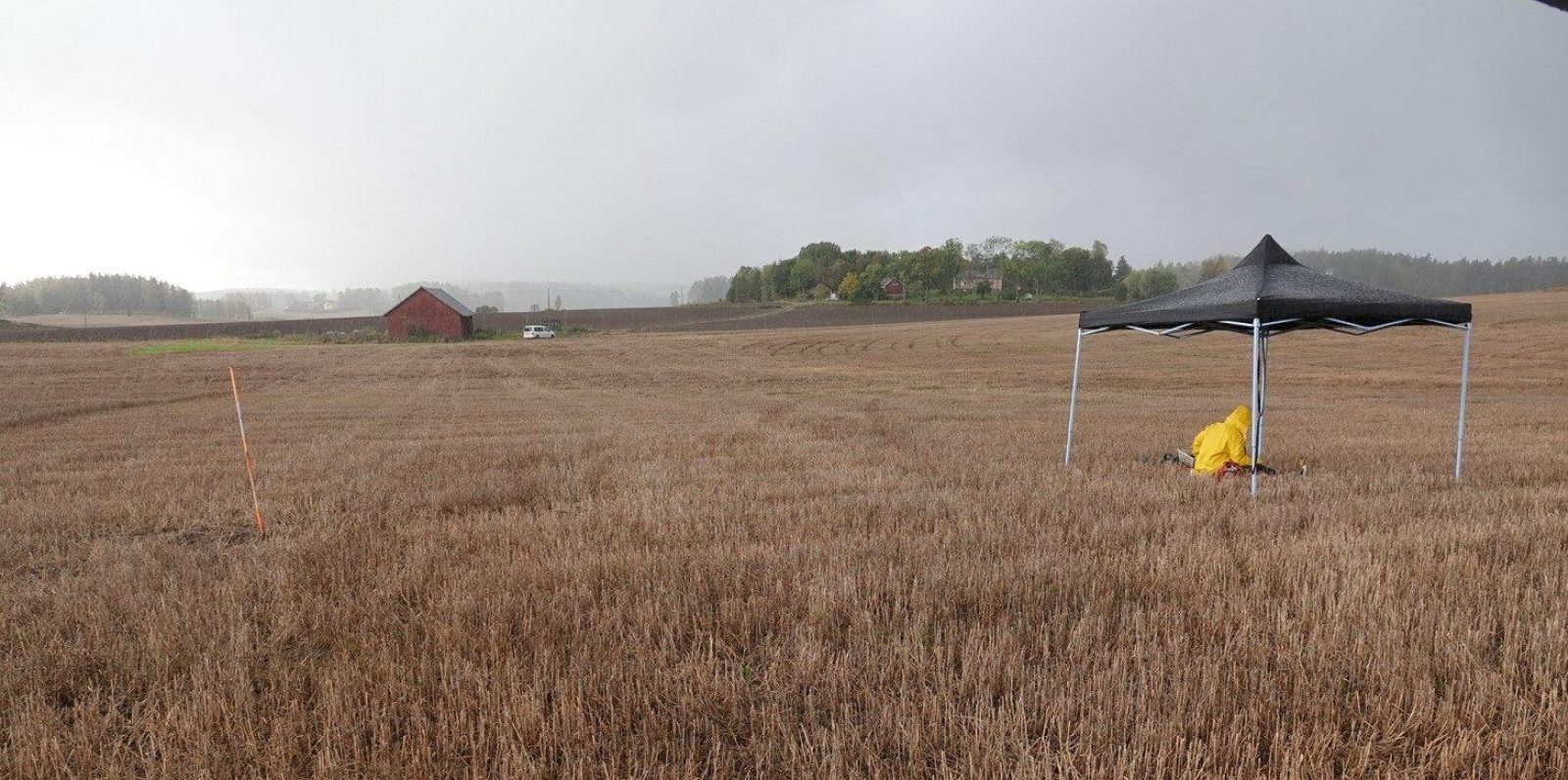Avaraa peltomaisemaa. Pellon keskellä on kevyt siirrettävä katos, jonka alla henkilö keltaisessa takissa.. Kuvaaja: Petro Pesonen