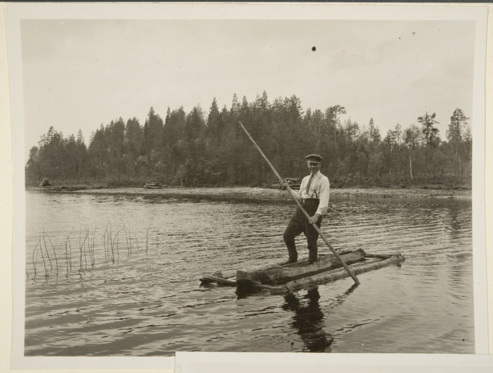Mustavalkokuvassa mies seisoo kolmiosaisen ruuhen kyydissä vedessä sauva kädessään. Taustalla metsää.. Kuvaaja: Tyyni Vahter Mustavalkokuvassa mies seisoo kolmiosaisen ruuhen kyydissä vedessä sauva kädessään. Taustalla metsää.. Kuvaaja: Tyyni Vahter