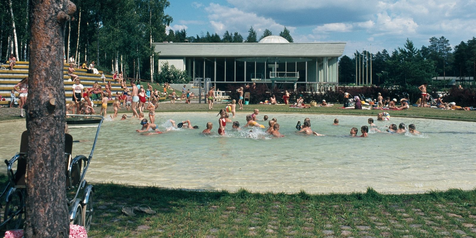 Lapsia uimassa ulkoaltaassa. Altaan ympärillä on lisää ihmisiä. Taustalla uimahallin päärakennus. Lapsia uimassa ulkoaltaassa. Altaan ympärillä on lisää ihmisiä. Taustalla uimahallin päärakennus.