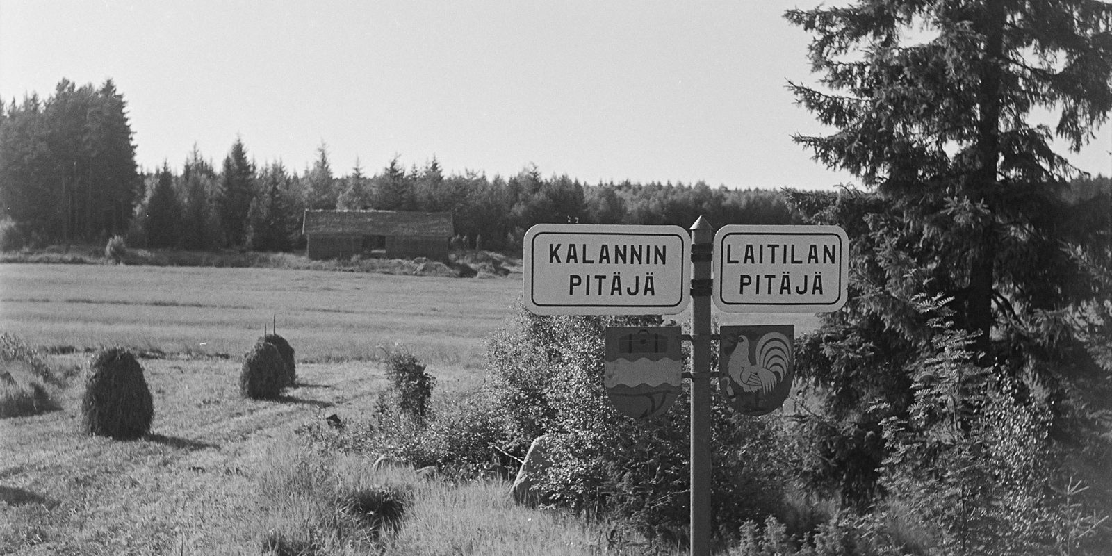 Mustavalkokuvassa peltomaisemaa ja etualalla kaksi tiekylttiä, joissa lukee Kalannin pitäjä ja Laitilan pitäjä.