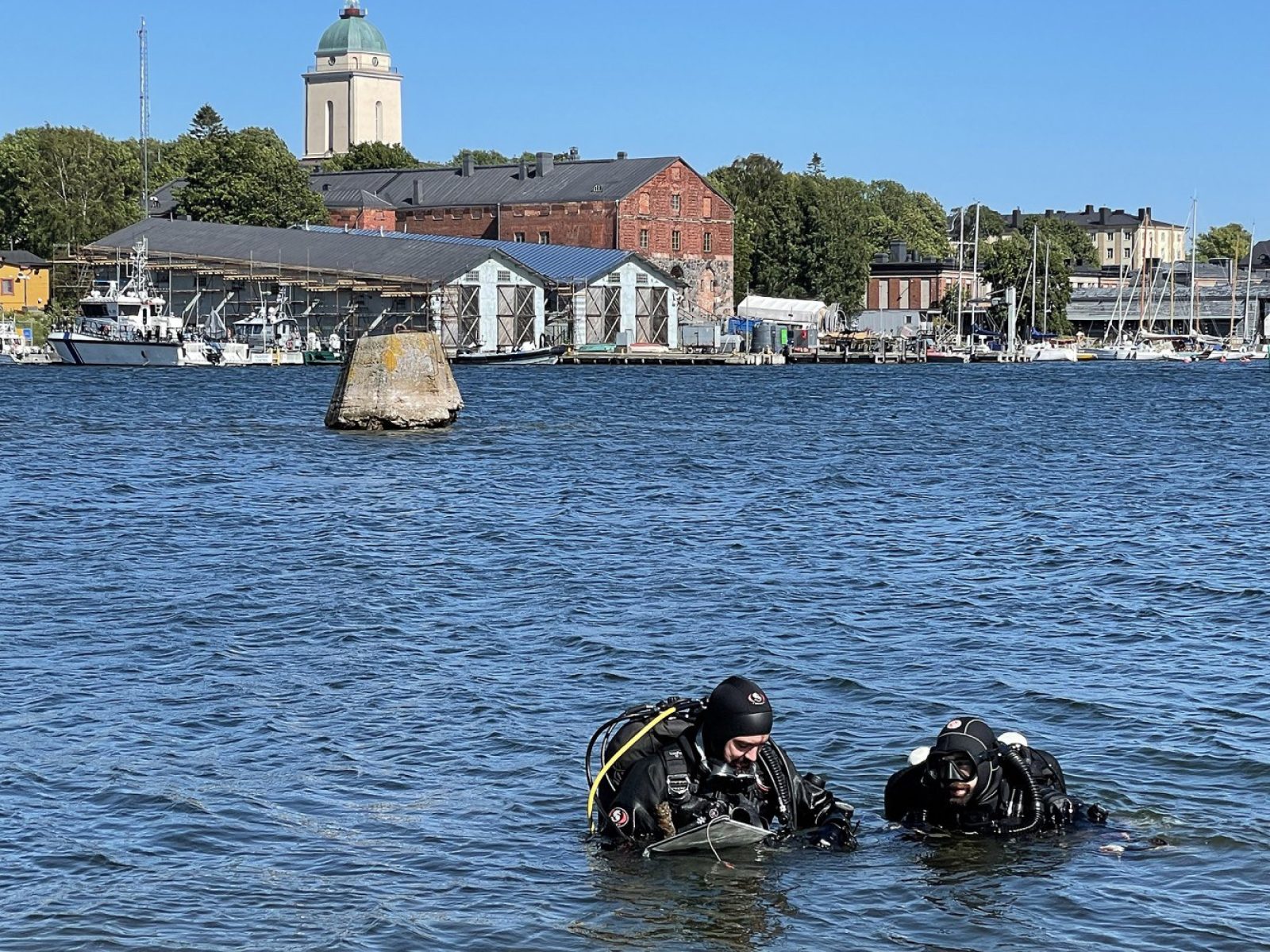 Kaksi sukeltajaa seisoo rintaa myöten vedessä. Taustalla Suomenlinnan rakennuksia.. Kuvaaja: Minna Koivikko, Museovirasto Kaksi sukeltajaa seisoo rintaa myöten vedessä. Taustalla Suomenlinnan rakennuksia.. Kuvaaja: Minna Koivikko, Museovirasto