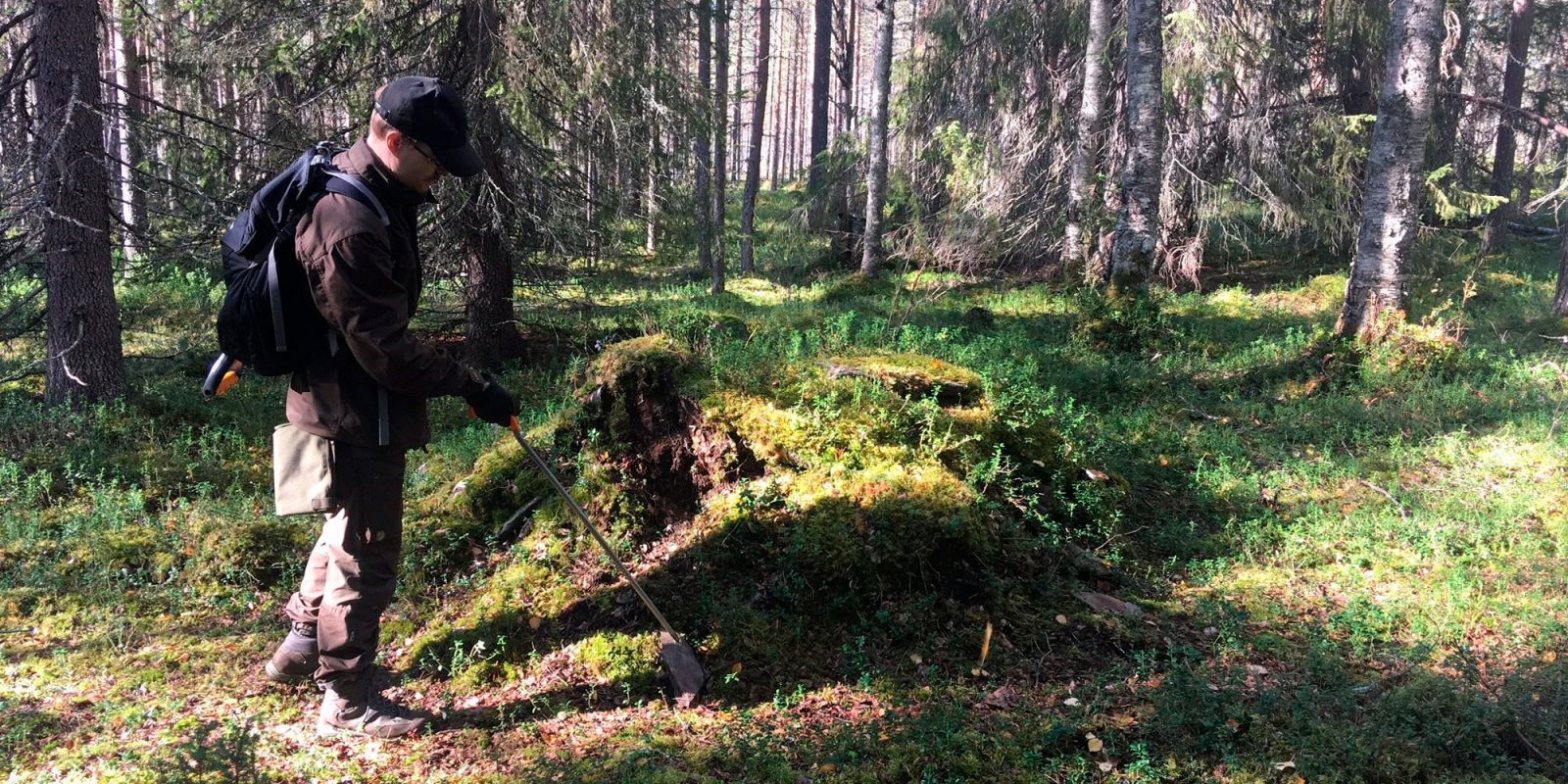 Arkeologi seisoo sammalen peittämän kivirakenteen vieressä.. Kuvaaja: Jouni Taivainen, Museovirasto. Arkeologi seisoo sammalen peittämän kivirakenteen vieressä.. Kuvaaja: Jouni Taivainen, Museovirasto.