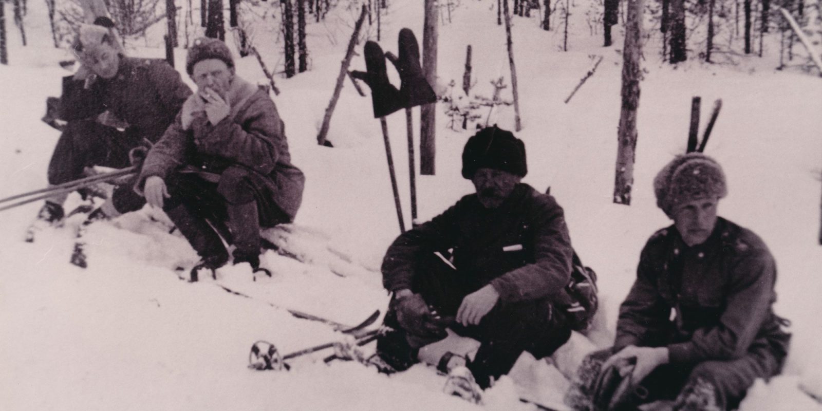 Mustavalkokuvassa neljä sotilasta hiihtovarusteineen istuu lumessa. Taustalla harvaa metsää. Mustavalkokuvassa neljä sotilasta hiihtovarusteineen istuu lumessa. Taustalla harvaa metsää.