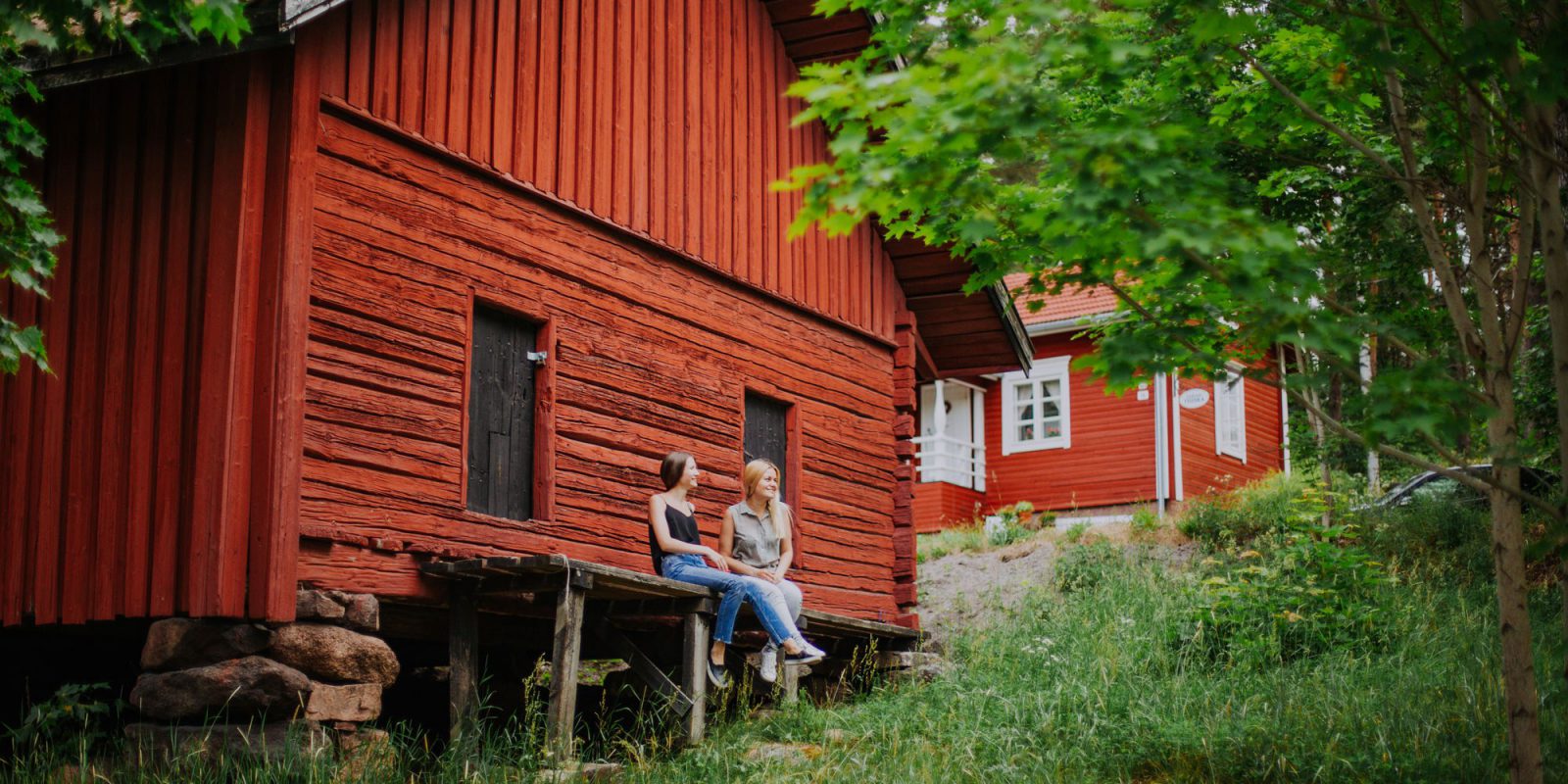 Kaksi naista istuu vanhan punaisen puurakennuksen reunustalla. Ympärlllä vehreitä puita, taustalla toinen punainen puutalo.. Kuvaaja: Julia Kivelä / Visit Finland. Kaksi naista istuu vanhan punaisen puurakennuksen reunustalla. Ympärlllä vehreitä puita, taustalla toinen punainen puutalo.. Kuvaaja: Julia Kivelä / Visit Finland.