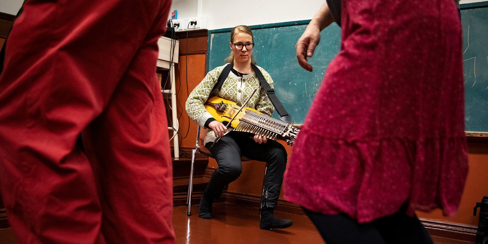 Avainviulunsoittaja istuu tuolilla liitutaulun edessä. Etualalla lähellä kamreaa kahden tanssijan jalat.. Kuvaaja: OKM / Emilia Anundi Avainviulunsoittaja istuu tuolilla liitutaulun edessä. Etualalla lähellä kamreaa kahden tanssijan jalat.. Kuvaaja: OKM / Emilia Anundi
