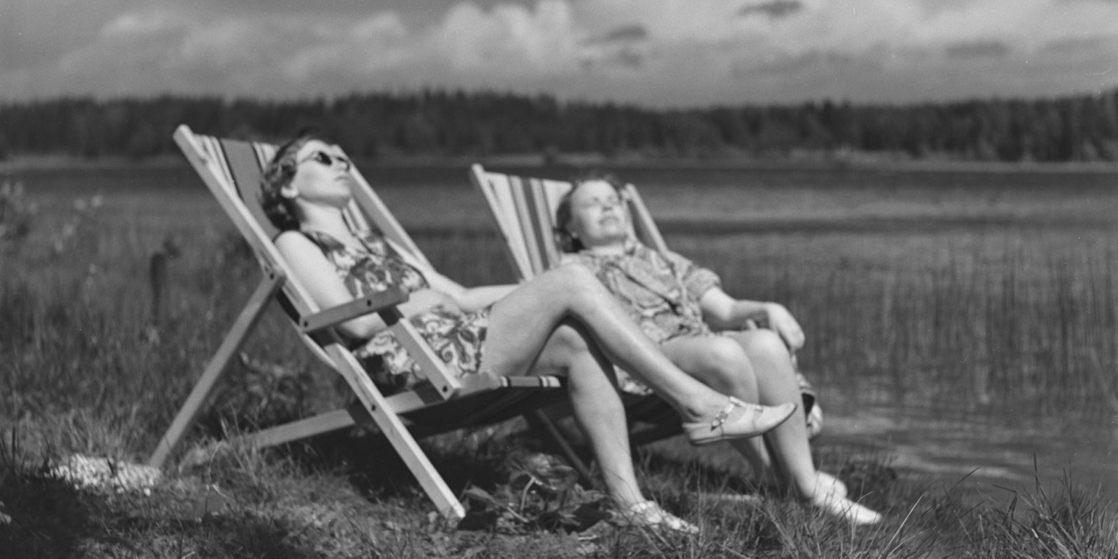 Mustavalkokuvassa kaksi naista aurinkotuoleissa pellon laidalla. Mustavalkokuvassa kaksi naista aurinkotuoleissa pellon laidalla.