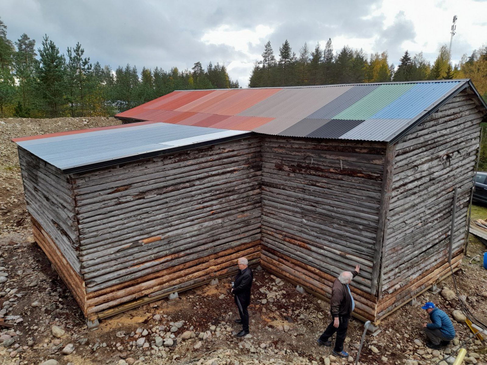 Vanha hirsirakennus, jonka peltikatto koostuu useista erivärisistä paloista. Rakennuksen juurella kolme miestä.. Kuvaaja: Marja Lähteenmäki / Lahden museot Vanha hirsirakennus, jonka peltikatto koostuu useista erivärisistä paloista. Rakennuksen juurella kolme miestä.. Kuvaaja: Marja Lähteenmäki / Lahden museot