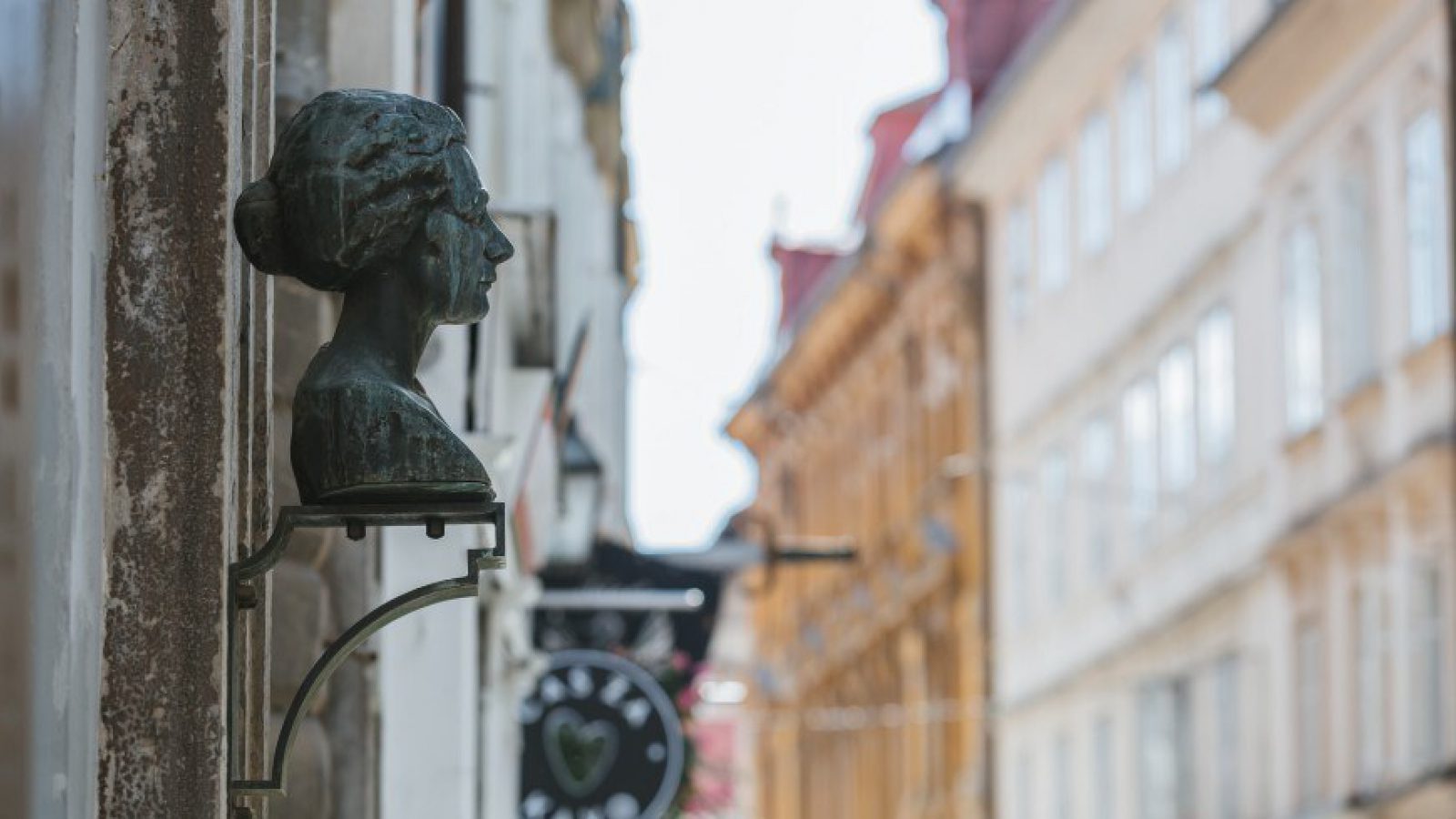 Pronssinen naisen rintakuva kiinnitettynä talon seinään. Taustalla epätarkkana vanhoja kerrostaloja. Pronssinen naisen rintakuva kiinnitettynä talon seinään. Taustalla epätarkkana vanhoja kerrostaloja.
